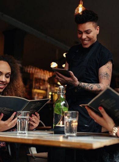 Young women seated at a table holding menus in a restaurant. Young girls at cafe making choice and place their order, while waiting standing by with a digital tablet.